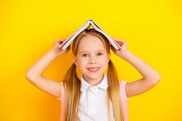 Cheerful schoolgirl holding an open book over her head against a bright yellow background,...