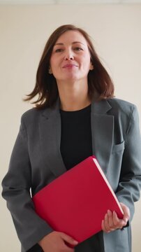 Confident businesswoman in gray suit holding red folder walking in office hallway with soft smile, ready for meeting or presentation, showing professionalism and corporate elegance