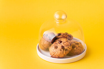 Stack of fresh chocolate American style chip cookies with nuts and chocolate drops on a tray with transparent lid on yellow background. Cookies with peanut butter filling inside a glass jar