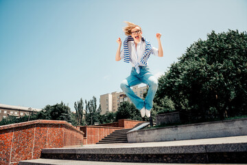 Joyful young woman jumping outdoor in a stylish striped shirt and jeans on a sunny day in an urban environment