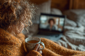 Young woman wrapped in blanket with tea attends virtual therapy session at home desk