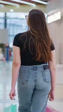 Behind view of young woman walking in mall with shopping bag, phone in back pocket, turning to look curiously behind while people pass by, vibrant mall atmosphere and modern retail shops
