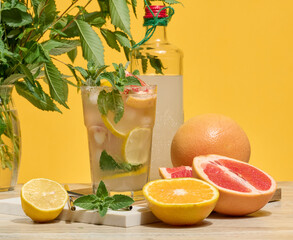 Fresh citrus drink with lemon, grapefruit, and mint in a glass with ice cubes and a straw. Surrounded by fresh citrus fruits and a bottle of homemade lemonade. Bright yellow background