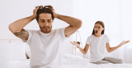 Sad millennial european husband with stubble sits on bed and holds head with hand, wife swears and...