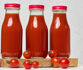 Glass bottles with tomato juice with red caps. Fresh red tomatoes lie on a wooden stand and in front of them, white background