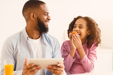 Time For Breakfast. Positive black girl eating sweet muffin cake looking at her smiling father who using digital tablet. Loving family sitting at dinner table, having meal together in the morning