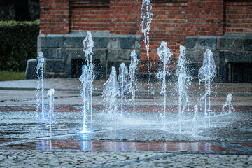 Jets of water shoot up from a ground fountain in front of a brick wall, lit by glowing blue lights, creating a dynamic and refreshing urban scene.
