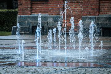 Jets of water shoot up from a ground fountain in front of a brick wall, lit by glowing blue lights, creating a dynamic and refreshing urban scene.