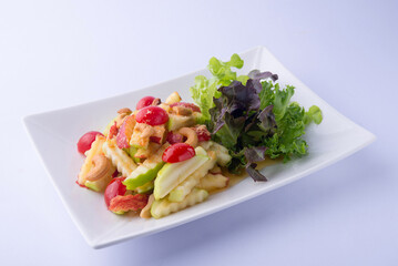Fruit salad: green and red apples, tomatoes, and cashews mixed with a spicy dressing, accompanied by green salad on a white plate, white background.