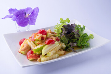 Fruit salad: Green and red apples, tomatoes, and cashews mixed with a spicy dressing, accompanied by green salad greens on a white plate and a white backdrop.