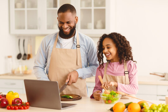 Portrait of happy African American man and little girl cooking in the kitchen and watching food video blog on laptop or reading recipe online using computer, cutting vegetables, preparing dinner