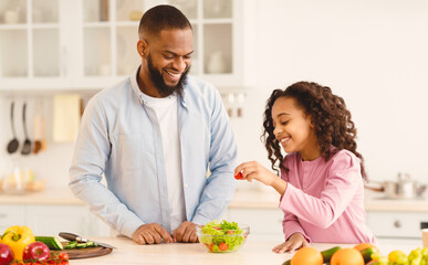 Portrait of happy african american father and little daughter cooking vegetable salad at modern kitchen, adding fresh tomato to bowl laughing together while making healthy lunch or breakfast at home
