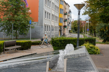 Modern urban fountain with water jets in city park with cyclist riding past colorful residential buildings and green landscaping. Concept of sustainable city living, bike-friendly infrastructure.