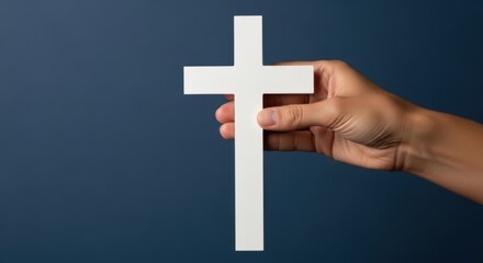 A hand holding a white Christian cross against a dark blue background provides a religious symbol. Faith and spiritual concept representation for Easter holiday.