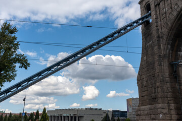 Low angle view of historic stone bridge tower with blue steel suspension cables against dramatic cloudy sky and modern city buildings. Concept of engineering marvel, urban architecture and heritage.