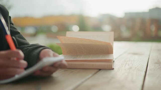 Close up of hand writing in notebook with pen beside open book on wooden table outdoors, book pages moving gently in wind, study moment in natural environment with soft blurred background