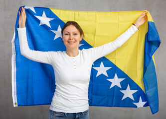 Happy young woman with large flag of Bosnia and Herzegovina posing gladly against light unicoloured...