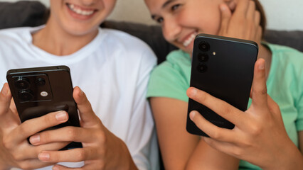 Two smiling teenagers sitting together, holding smartphones and enjoying time on social media. Close-up of hands and phones with blurred happy faces in the background.