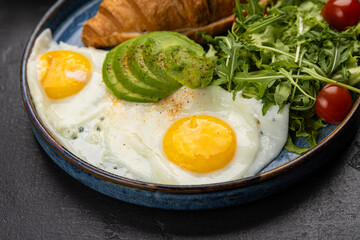 Healthy breakfast with croissant, avocado, sunny side eggs and salad on dark background