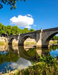 Fototapeta premium Ancient stone arch bridge over a calm river, reflected in the water, with lush green trees and a bright sky