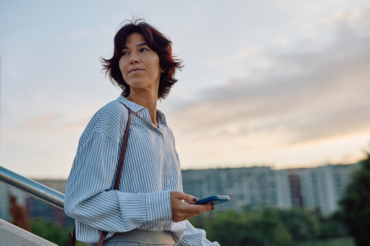 Businesswoman walking up stairs holding smartphone and looking away with buildings and trees in background at sunset