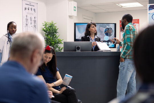 Friendly female receptionist with clipboard greets african american male visitor at hospital front desk. Clinic staff hands over medical form to black man for scheduled checkup in healthcare facility.