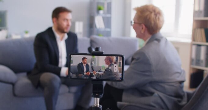 Female host of business media content channel interviews young male influencer sharing professional success story. Blur background soft focus closeup screen of modern cell phone device recording video