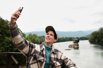 Obraz premium Woman taking a cheerful selfie by the river in front of the famous Drina house on the rock in Serbia. Female travel blogger
