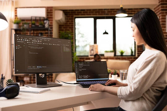 Focused female developer programming on laptop in home office. Asian woman building software and coding website remotely from modern apartment using digital tools for web development.