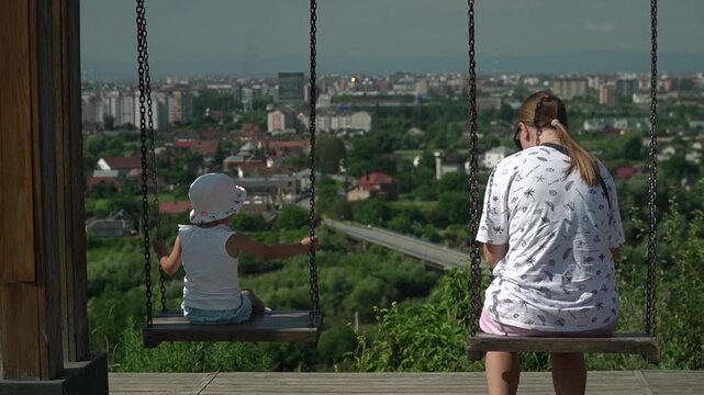 A girl and her mother enjoy the view of the city from a hill while riding a swing, looking at everything, the child points, a bright sunny summer day, slow motion
