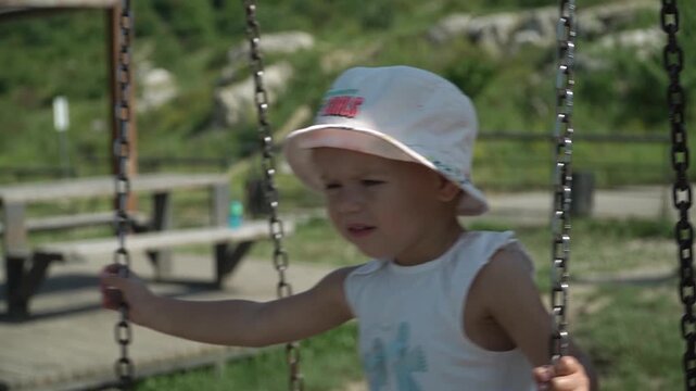 A girl in a light-colored Panama hat swings on a swing, holding onto the chains, pointing forward and intently examining everything around her, on a bright summer day