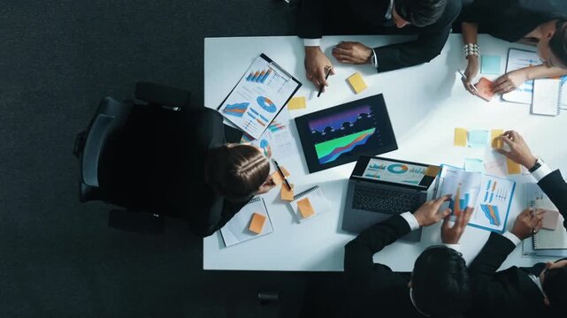 Top down aerial view of diverse business people discuss and brainstorm marketing idea while looking at financial graph at table with marketing document and sticky notes scatter around. Directorate.