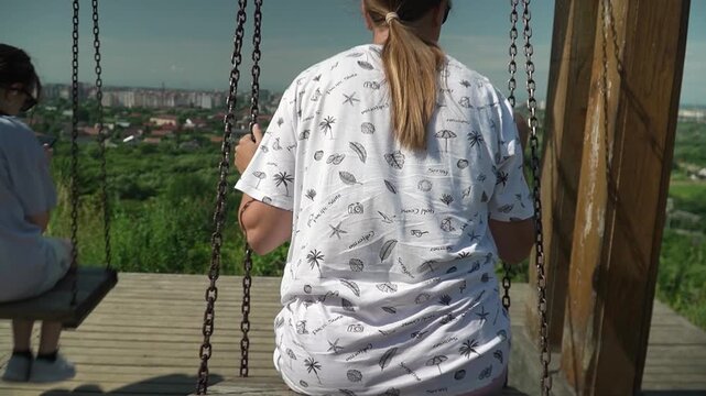 A girl with long blond hair pulled back into a ponytail, wearing a white T-shirt, riding a swing with a panoramic view of the city, on a bright sunny summer day, slow motion