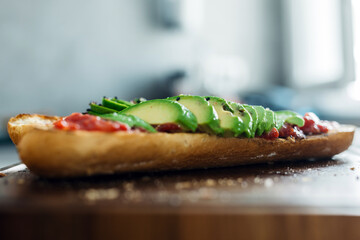 Freshly prepared sandwich with sliced avocado, tomato sauce, and herbs on a wooden cutting board, showcasing vibrant colors and appetizing textures in a modern kitchen setting