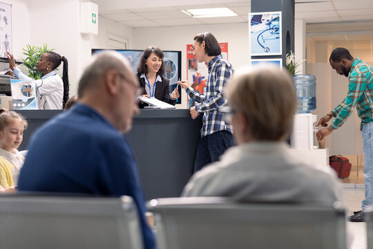 Receptionist at front desk supports patient intake by updating health records and scheduling treatment visits in busy clinic. Hospital staff discussing upcoming medical checkups with asian man. - Powered by Adobe