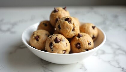 Several raw chocolate chip cookie dough balls piled high in a white bowl on a marble surface. These sweet treats look soft and chewy ready for baking.
