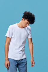 Fototapeta premium Young man with curly hair wearing white t-shirt and blue jeans stands looking down thoughtfully against bright blue background in casual style.