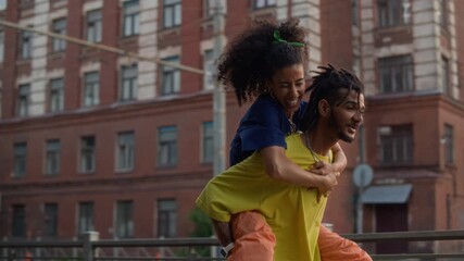 Medium slowmo shot of carefree young biracial girl jumping on her boyfriends back and running together in city street on beautiful summer day, sharing happy romantic moments - Powered by Adobe