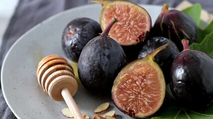 A rustic still life of fresh Black Mission figs, some whole and some cut in half to reveal their vibrant red flesh. The figs are arranged on a gray ceramic plate with a wooden honey dipper and chopped