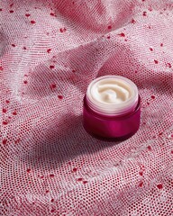 Elegant pink cosmetic cream jar on textured fabric with red heart pattern.