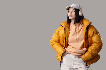 Young woman in stylish puffer jacket listening to music on grey background