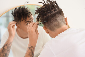 Fototapeta premium Young man with dreadlocks combing hair near mirror in bathroom, back view