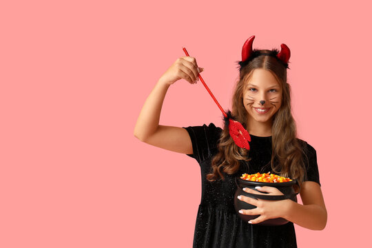 Cute happy girl dressed as demon for Halloween with pot of candy corns and trident on pink background