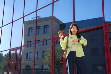 Young Asian woman with coffee looking at wristwatch outdoors. Time management concept