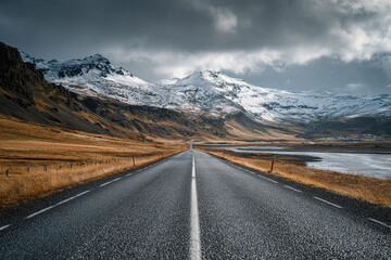 A long straight road stretches through a mountainous landscape with snow-capped peaks under a dramatic cloudy sky.