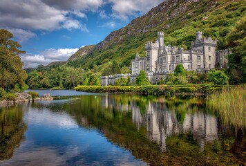 Historic gray castle reflected in a tranquil lake beneath lush green hills under a bright blue sky with scattered white clouds