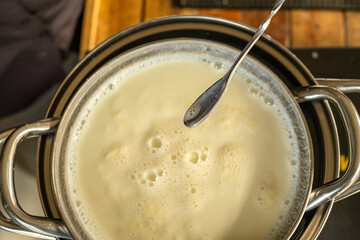 Close-up top view of boiling fresh cow milk in a stainless steel pot with foam texture and a spoon above, showing natural dairy preparation in a warm kitchen atmosphere.