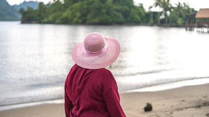 A woman wearing a pink hat and red outfit stands on the sandy beach, looking at the calm sea and distant green island in the afternoon.