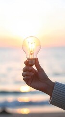 A hand holds a light bulb against a blurred seascape with a warm sunset. Light shines through bulb, creating a hopeful atmosphere