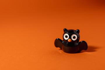 Close-up of a small owl figurine with large eyes casting a dramatic bat-shaped shadow against an orange background. The playful Halloween-inspired setup creates a spooky yet cute atmosphere